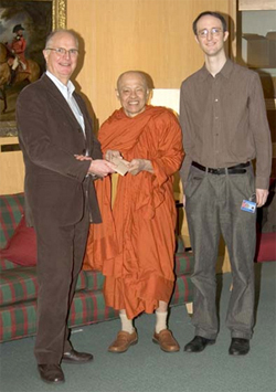 Three men standing posed for a photograph: two in formal western clothes, one in orange monastic robes. 