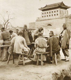 Men in padded jackets sitting around a table, outside a thick city wall's checkpoint gate. Historical sepia photograph. 