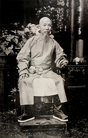 A man in Chinese official's robes with shaven head and beard, seated in a space with ornately carved wooden screens. 