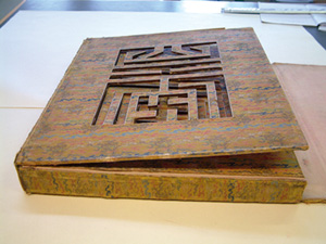 Rectangular box designed to cover a book, with a woven pattern, and a geometric design cut out of the front cover flap.