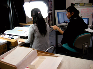 Colleagues seated at work benches, one looking at a document and one at its digitisation on a screen. 