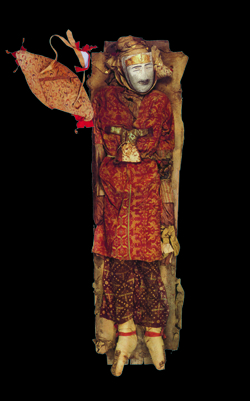 A body wearing a white mask and highly patterned funerary clothes.