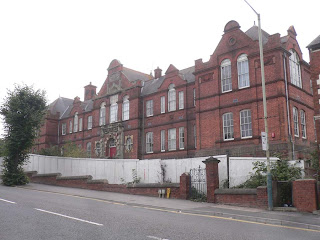 Image of a red brick Victorian style school building with a white hoarding outside.