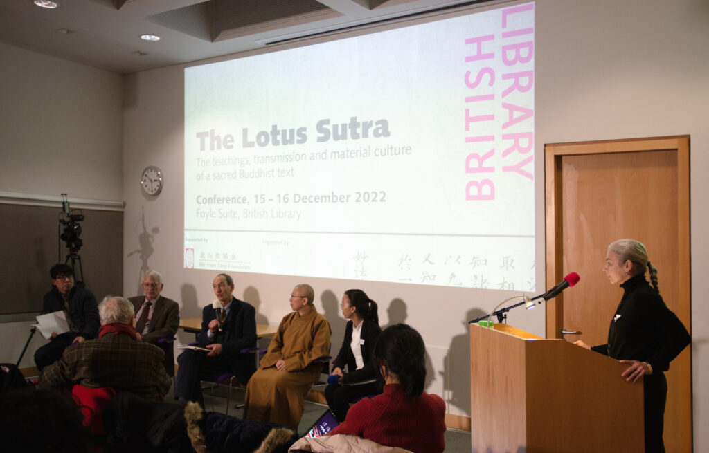 A group of people sitting in front of a presentation screen which projects the title of the Lotus Sutra conference. One holds a microphone and seems to be answering a question. 
