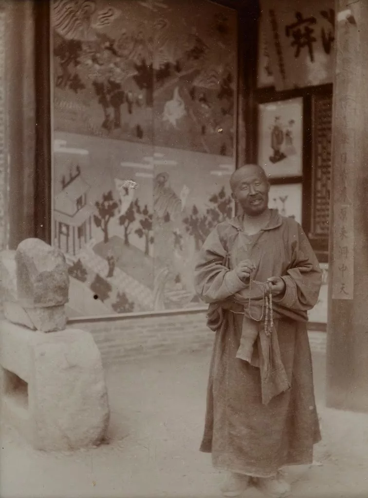 Man in monk's robes with rosary standing in front of a painting. Historic sepia photograph. 