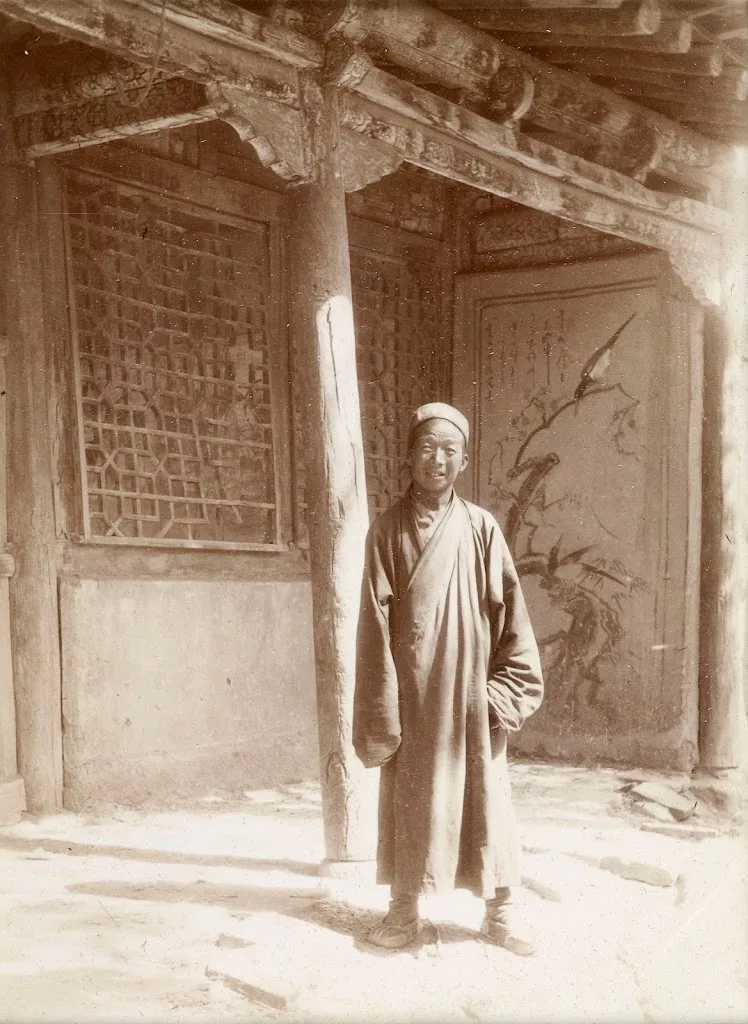 A man standing by a pillar, on the verandah of a building with latticework on the windows, with a painting behind him. Historic sepia photograph. 