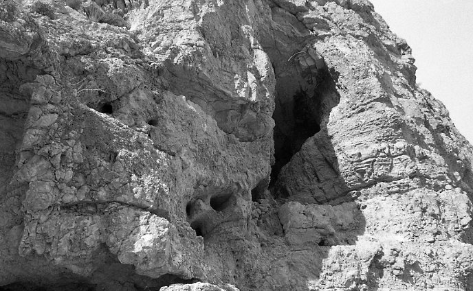 A black and white photograph of a cave entrance in a cliff face. 