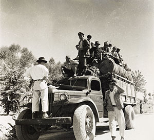Several people standing on the raised and covered bed of a mid-twentieth century truck. Black and white historical photograph.