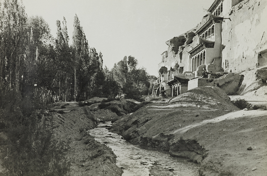 Landscape image showing trees, a river, and caves with terrace balconies built into a cliff face. Historical black and white photograph.