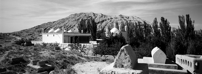 Modern black and white photograph of a stone mausoleum, with a building with round domes behind and a mountain in the distance.