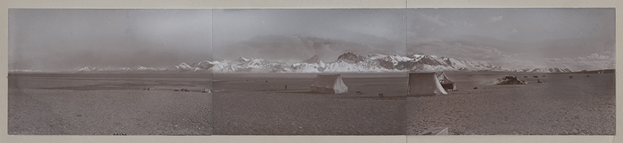 Panoramic image, black and white historic photograph, showing flat desert with a mountain range in the distance. 