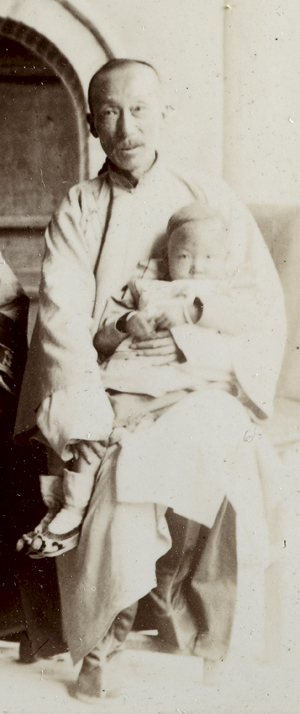 A robed and moustached Chinese man, sitting, with a child in his arms. Historical black and white photograph. 