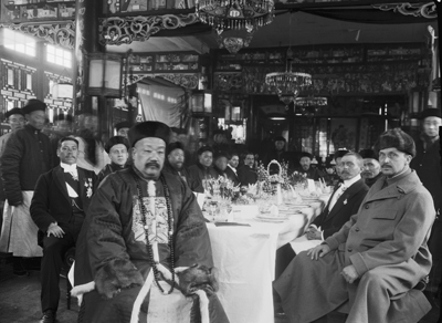 Two men, one in ceremonially embroidered Chinese official robes and one in a heave winter coat and fur hat, sitting at the head of a banqueting table. Historical black and white photograph.