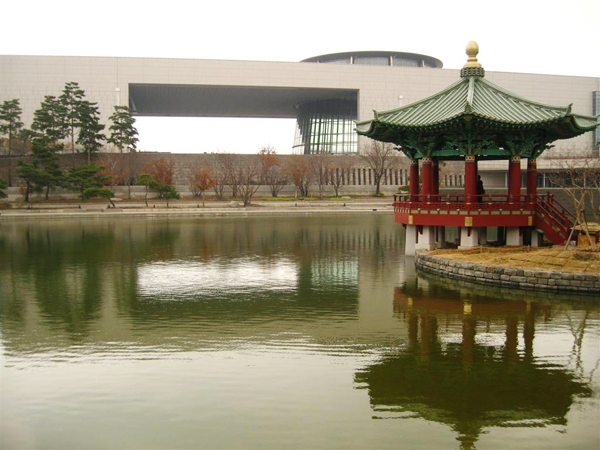 A traditional Korean roofed building in an artfical lake in front of a modern museum building. 