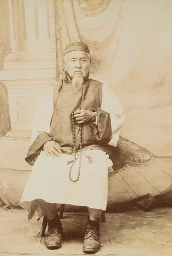 Sepia photograph of a man with a beard, seated, holding a length of prayer beads. 
