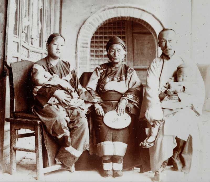 A young woman, old woman and man sitting together. The young woman and man each have a child in their arms. Black and white historical photograph. 