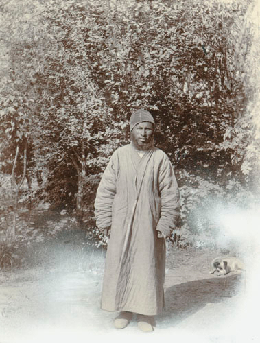 A man in a thick robe and turban standing by a tree, with a small dog lying down behind him.