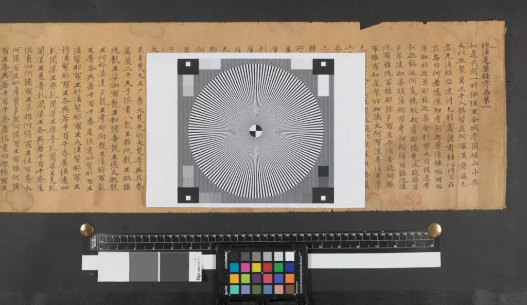 A scroll laid out flat to be digitised, with a black and white patterned card in front and a colour chart below.